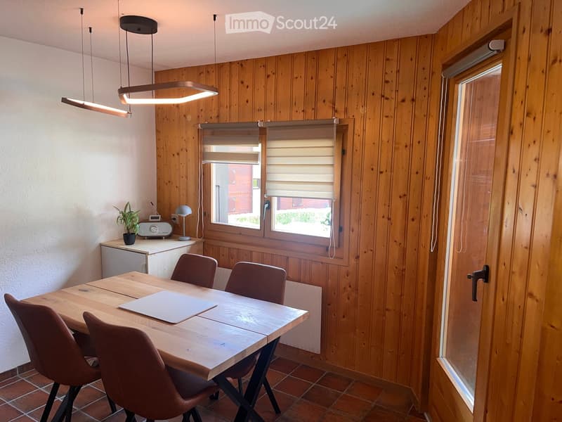 Dining area with wooden walls, tiled floor, and large windows with blinds. Includes a wooden dining table with four chairs, a small plant, a radio, and a lamp on a sideboard.