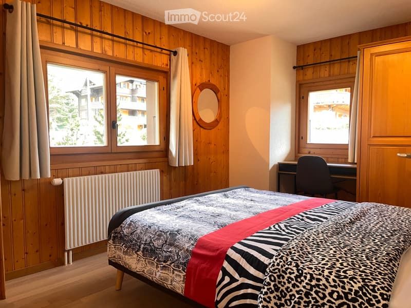 Cozy bedroom with wooden walls, large windows, a bed with a patterned duvet, a desk with a chair, and a round mirror on the wall.