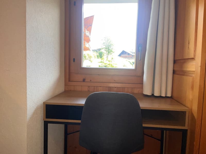 A small, furnished room with a wooden desk, a single chair, and a window with curtains offering a view of the outside.