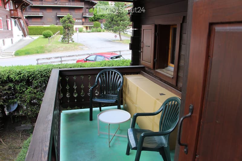 Balcony with green plastic chairs, a small round table, and a cardboard box, overlooking a street with a red car and surrounding greenery.