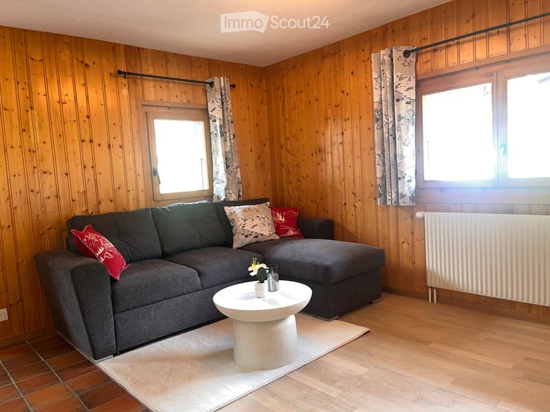 Living room with wooden walls and flooring, grey sectional sofa with decorative pillows, white coffee table with a small flower vase, radiator on the wall, and large windows with patterned curtains.