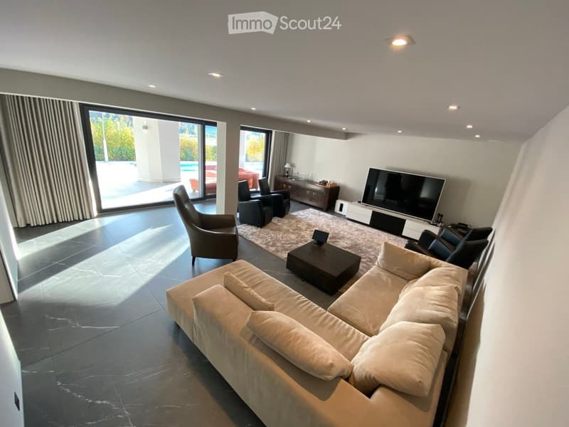 Modern living room with large windows, beige sofa, coffee table, armchairs, TV, and side table.