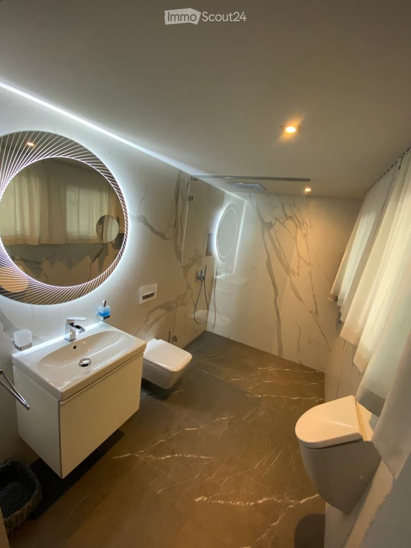 Modern bathroom with marble flooring and walls, featuring a large round mirror, a sink with a modern faucet, and a toilet.