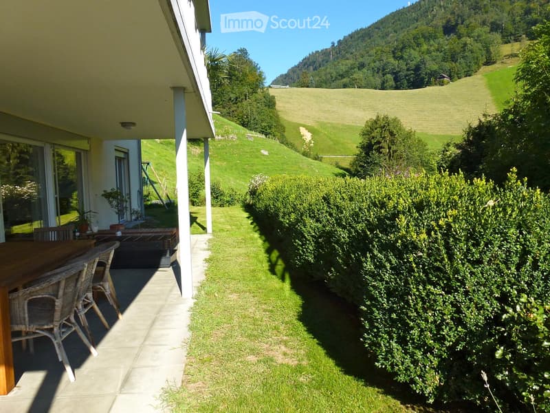 Outdoor patio with wooden table and wicker chairs, surrounded by green lawn and bushes, with a scenic mountain view in the background.