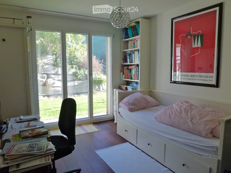 Bright bedroom with a single bed, a bookshelf, a chair, and a large window with a view of the garden.