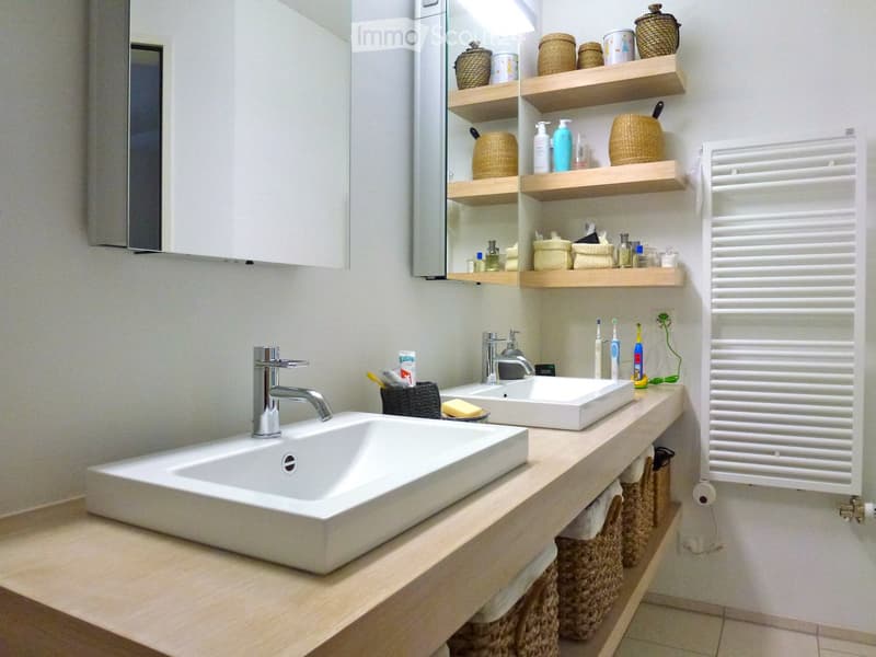Modern bathroom with two white sinks, wooden countertop, shelves with decorative items, and a white radiator.