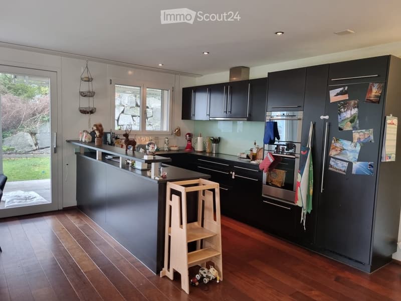 Modern kitchen with dark grey cabinets, stainless steel appliances, and a central island. The space features a wooden floor, large windows, and various kitchen items including a refrigerator, oven, and a small wooden stool.