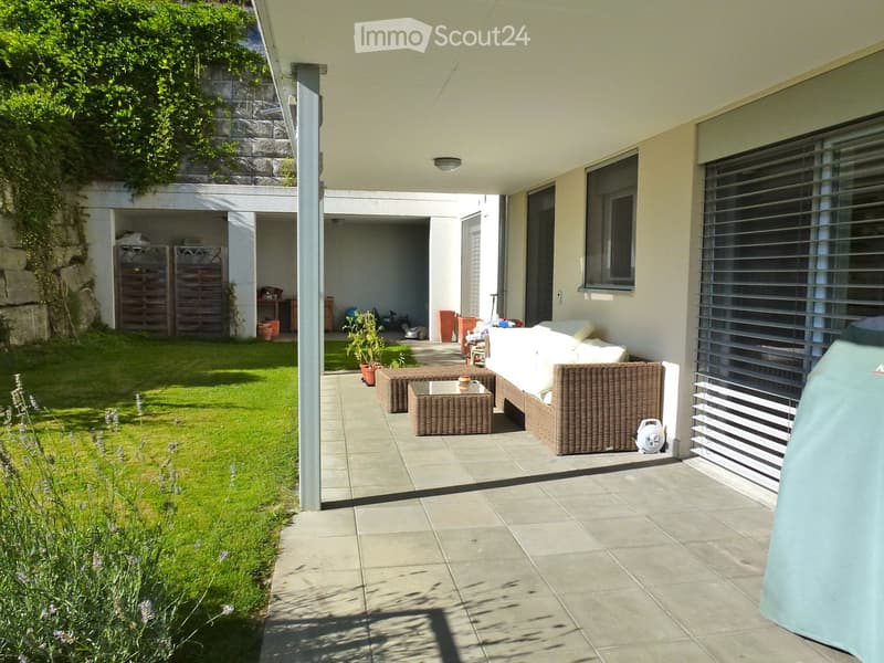 Outdoor patio with a sofa, wicker chairs, coffee table, potted plants, and a small fan. The area is tiled and has a grassy lawn with lavender plants on one side.