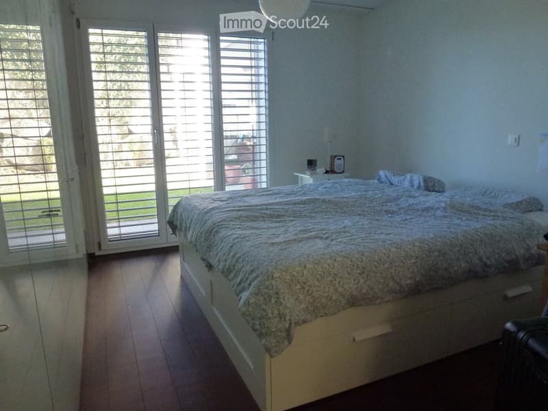 Bright bedroom with wooden flooring, large sliding glass doors leading to a garden, a neatly made bed with patterned bedding, and a small nightstand with a lamp.