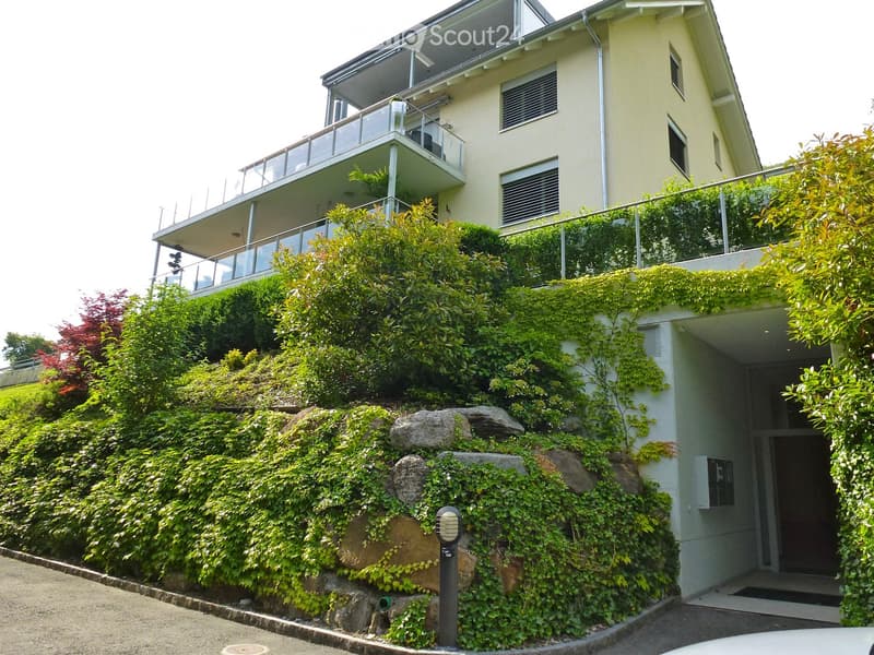 Modern two-story house with light yellow walls, large windows, and a balcony on the upper level. The house is surrounded by lush greenery and has a covered entrance area.