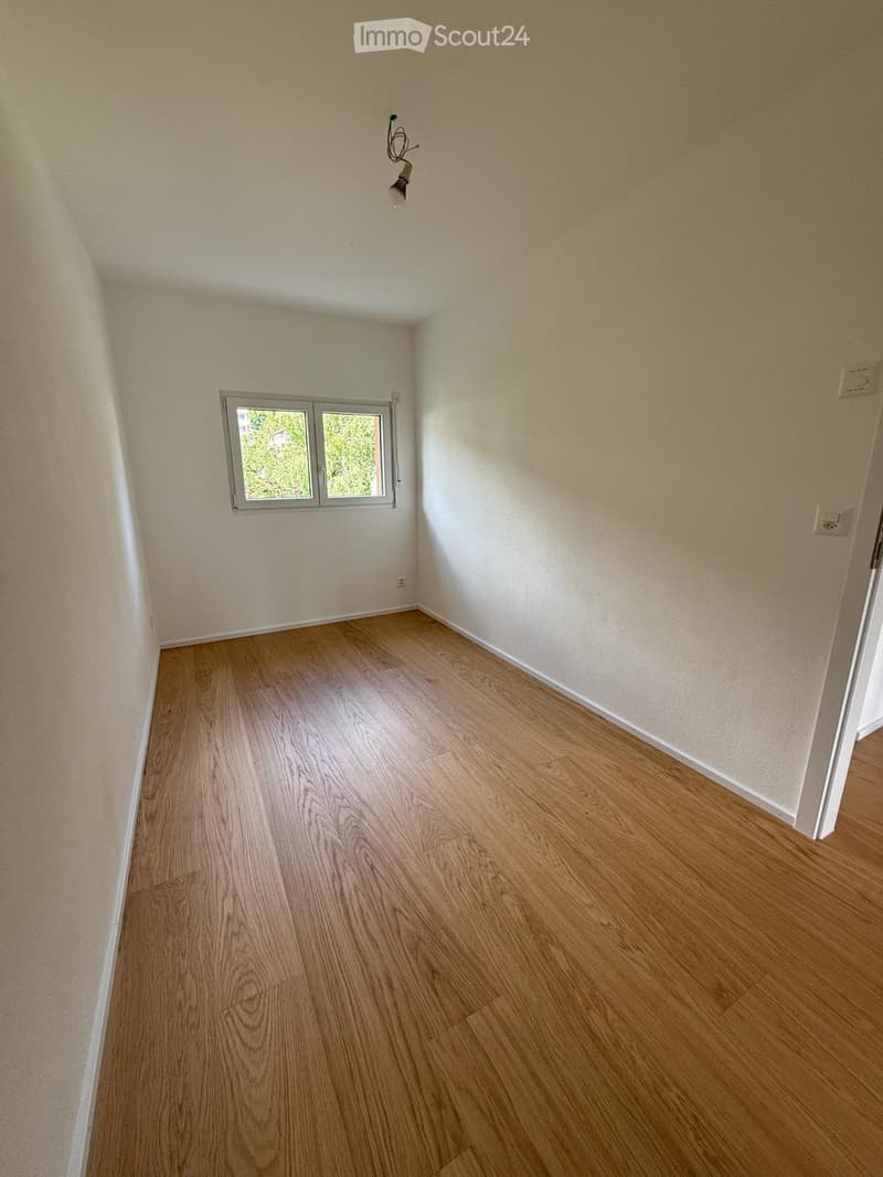 Empty room with light wooden flooring, white walls, and a small window with a view of greenery outside.