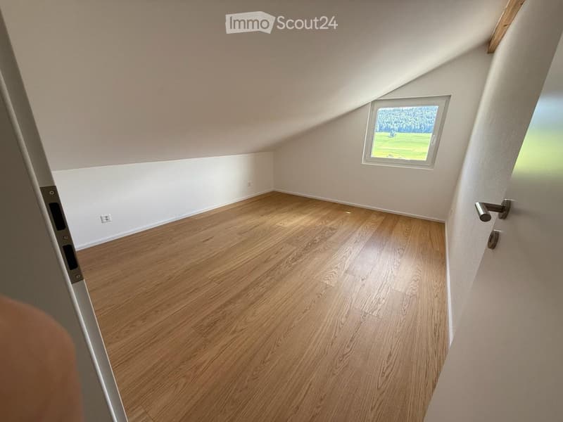 Empty attic room with wooden flooring, white walls, and a small window showing a green outdoor view.