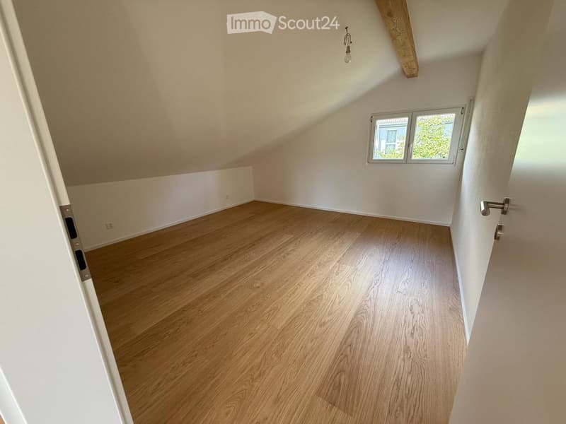 Empty attic room with wooden flooring, white walls, and a small window letting in natural light.