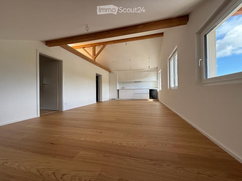 Empty room with wooden flooring, white walls, exposed wooden beams, and large windows allowing natural light. Kitchen area visible in the background with white cabinets and appliances.