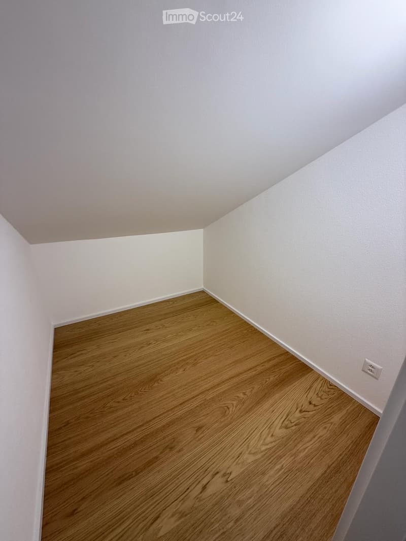 Empty room with light-colored walls and wooden flooring, showing a corner view with a visible electrical outlet on one wall.