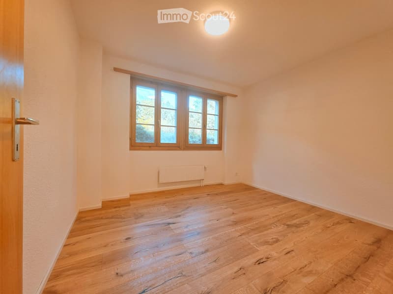 Empty room with wooden flooring, white walls, a large window with wooden frame, a radiator, and a round ceiling light fixture.