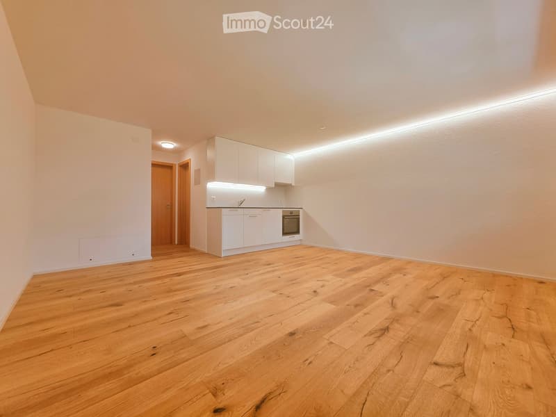 Empty room with light wooden flooring, white walls, and a small white kitchen area with cabinets and an oven. The space is well-lit with recessed lighting and has a door leading to another area.