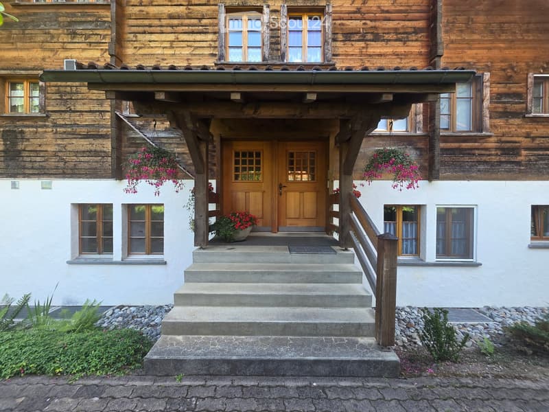A two-story wooden house with a covered entrance, stone steps, and hanging flower baskets.