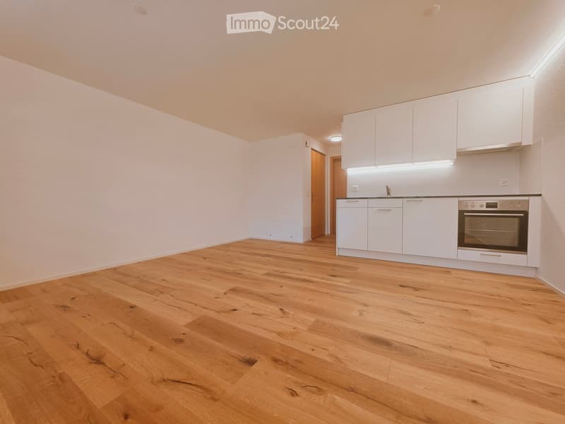 Empty room with light wooden flooring, white walls, and a small kitchen area with white cabinets and a built-in oven.