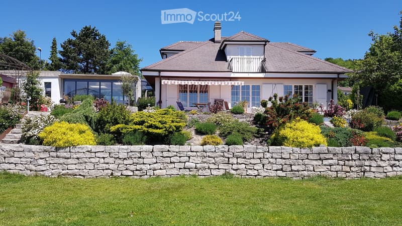 A well-maintained garden with a stone wall, lush greenery, and a two-story house with a balcony and patio area.