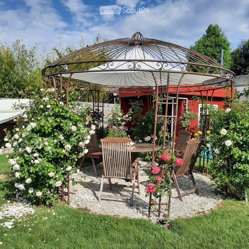 Garden area with a metal gazebo, wooden table, and chairs surrounded by flowers and greenery.