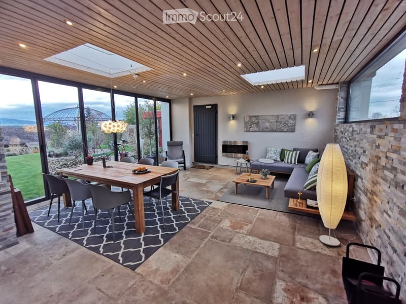 Open-plan living and dining area with large windows offering a scenic view, featuring a sofa, coffee table, dining table with chairs, fireplace, and decorative lighting.