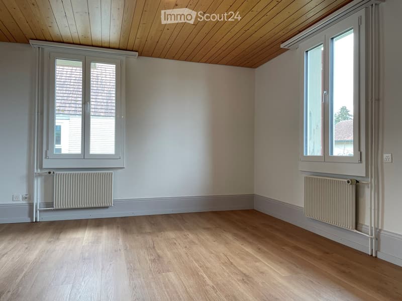 Empty room with wooden flooring, white walls, and large windows with white frames. Two radiators are visible on opposite walls.