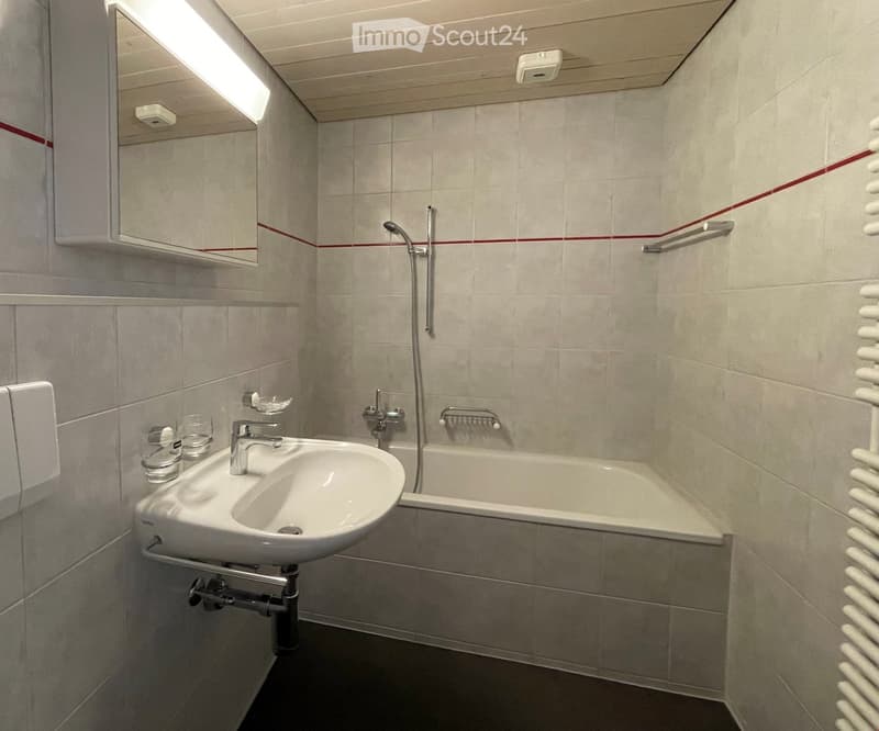 Modern bathroom with a white sink, bathtub, mirror, and towel rack. The walls are tiled in light gray, and the floor is dark.