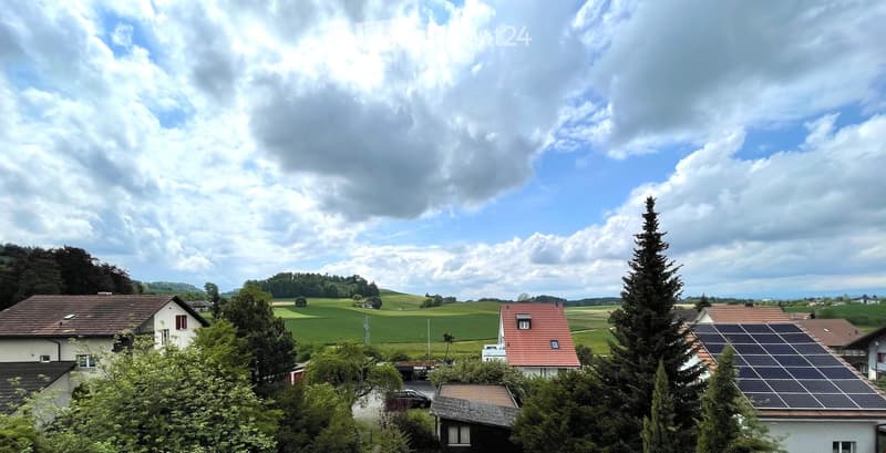 Residential area with multiple houses, green fields, trees, and a partly cloudy sky.