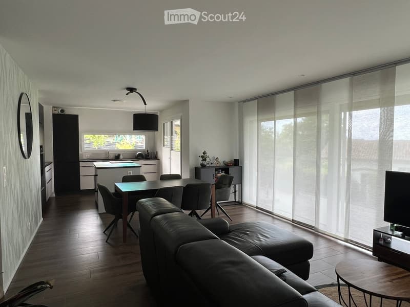 Modern living and dining area with a leather sofa, coffee table, dining table with chairs, kitchen island, and large windows with blinds.