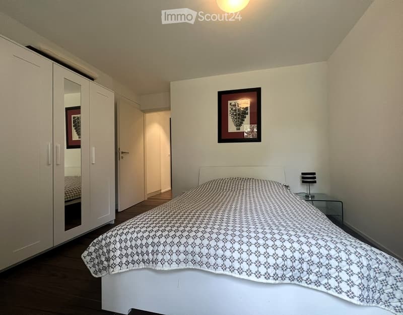 Bedroom with a white bed, patterned duvet, white wardrobe, nightstand with lamp, and picture frame on wall.