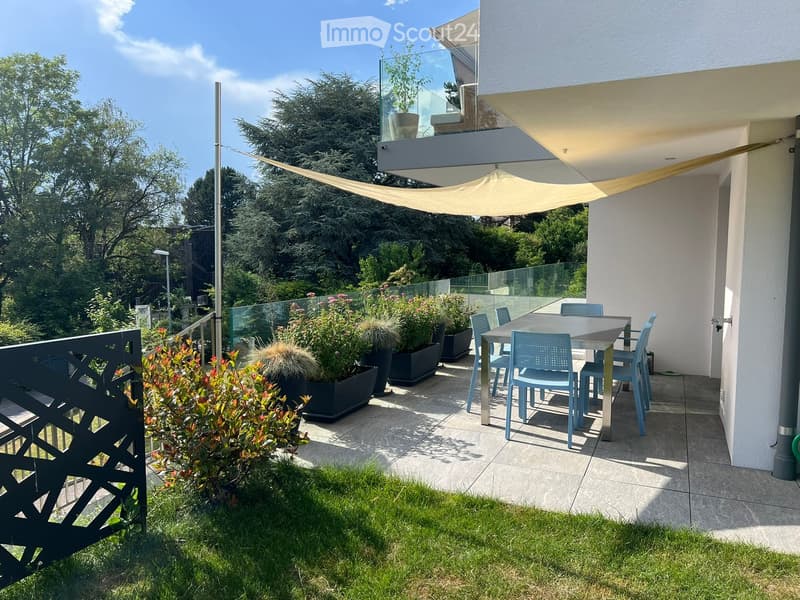 Balcony with a dining table and chairs, surrounded by potted plants and a sunshade, with a view of greenery.