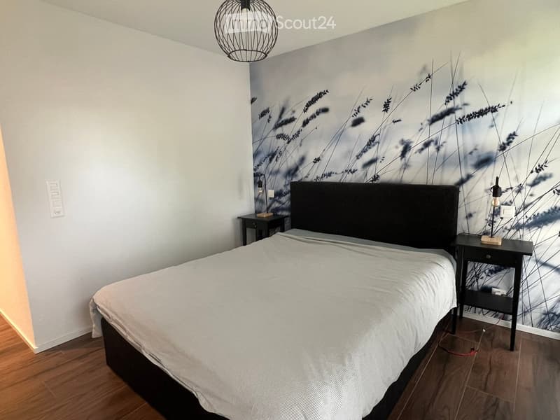 Modern bedroom with a large bed, nightstands, and a decorative wall mural featuring abstract plant designs. The room has wooden flooring and a hanging pendant light.