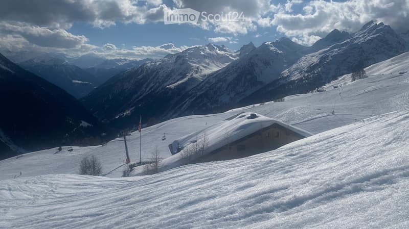A snowy mountain landscape with a small building partially covered in snow, surrounded by snow-covered slopes and distant peaks.
