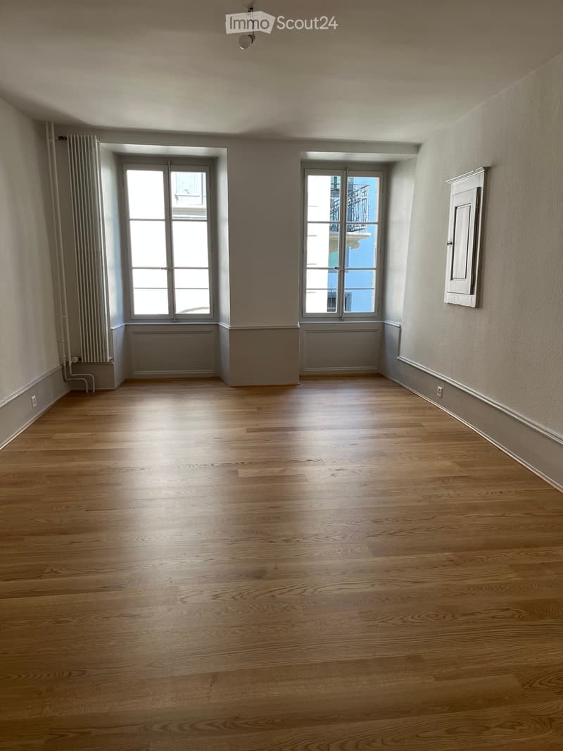 Empty room with wooden flooring, two windows, radiator, and a door frame.