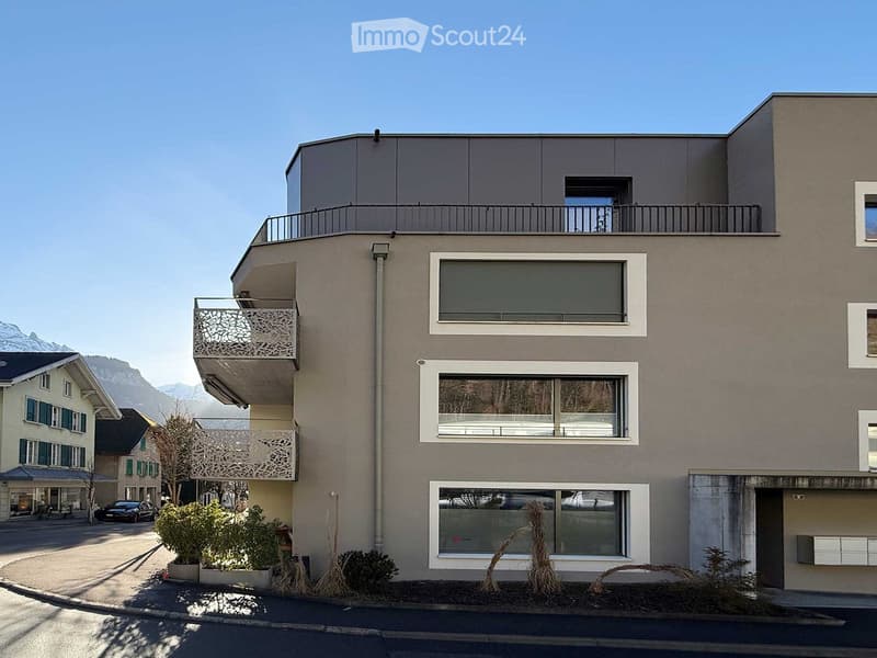 Modern apartment building, three stories, gray exterior, white windows, multiple balconies, fenced garden in front