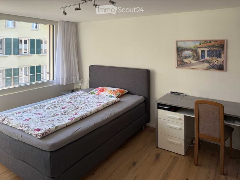 Single bed, headboard, floral comforter, nightstand with lamp, desk with chair, framed picture, curtains, wooden floor
