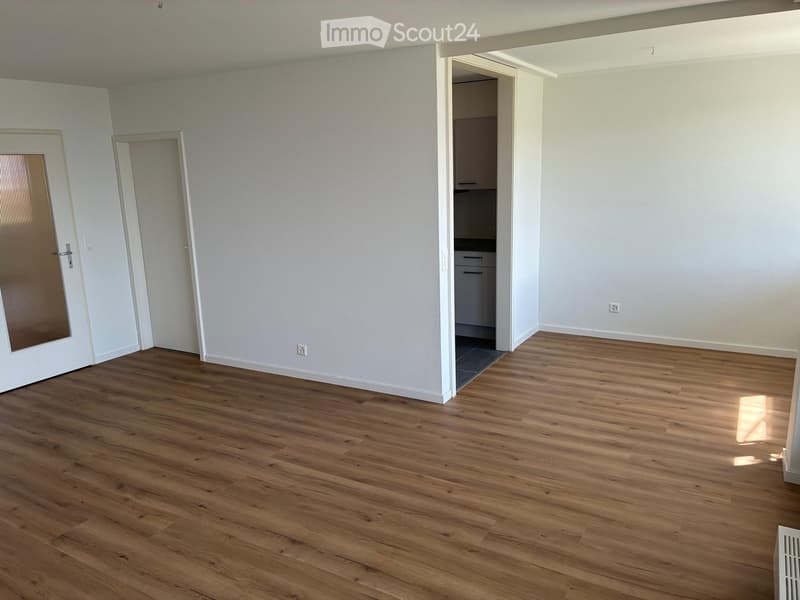Empty room with light-colored walls and wooden flooring, featuring a partially visible adjacent kitchen area with cabinets and drawers.