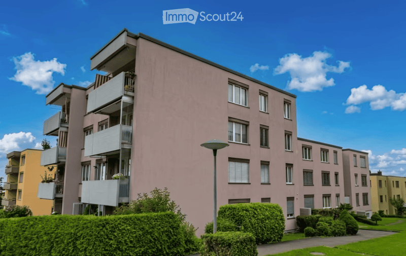 Modern multi-story residential building with pink facade, multiple balconies, and greenery in front.