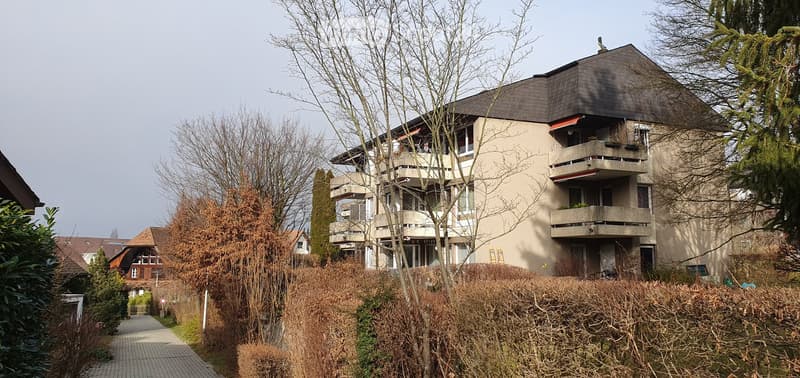 Modern multi-story apartment building with balconies, surrounded by trees and shrubs.