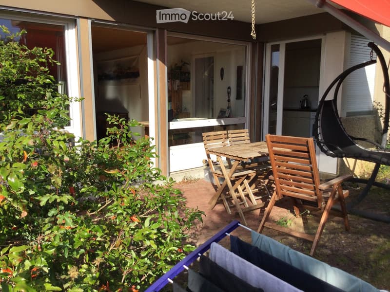 Outdoor patio with wooden table and chairs, clothesline with hanging towels, and a hanging egg chair. The patio is adjacent to a house with large glass doors and visible indoor furniture.