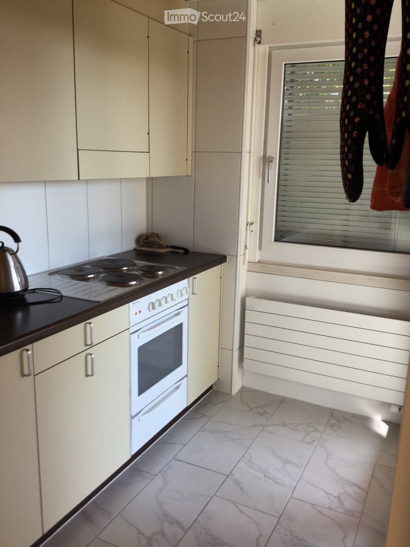 Modern kitchen with white cabinets, a black countertop, a white oven, and a stainless steel teapot on the stove. The floor is tiled with a marble-like pattern.