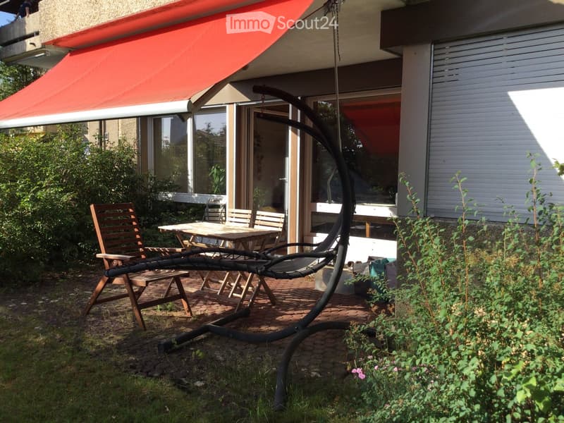 Garden area with a wooden table, chairs, a swing, and a red awning attached to the house.