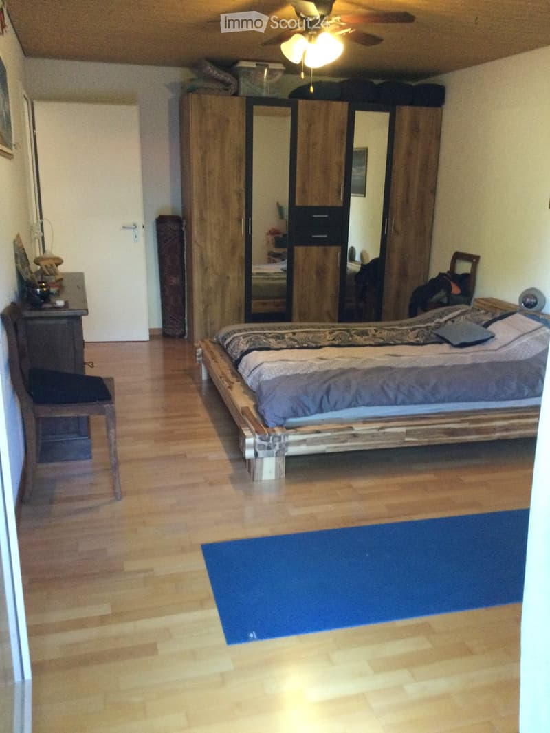 Cozy bedroom with wooden floor, large bed with patterned bedding, wardrobe with mirrored doors, small table with items, and a chair.