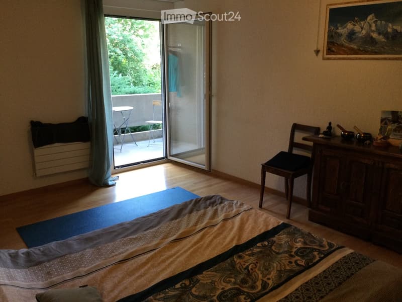 Bright bedroom with wooden flooring, a bed with patterned bedding, a yoga mat, a small table, and a chair near a large sliding glass door leading to a balcony.