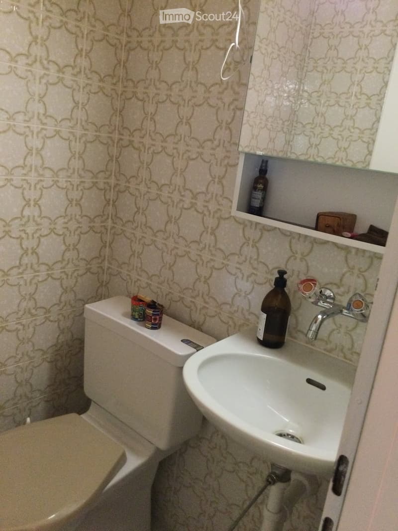Bathroom with a toilet, sink, and mirror. The sink has a bottle and a faucet, and the toilet has a few cups on top.