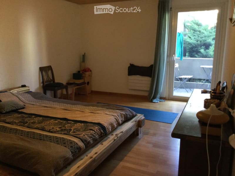 Cozy bedroom with wooden floor, large bed, chair, small table, and a balcony with outdoor furniture visible through the sliding glass door.
