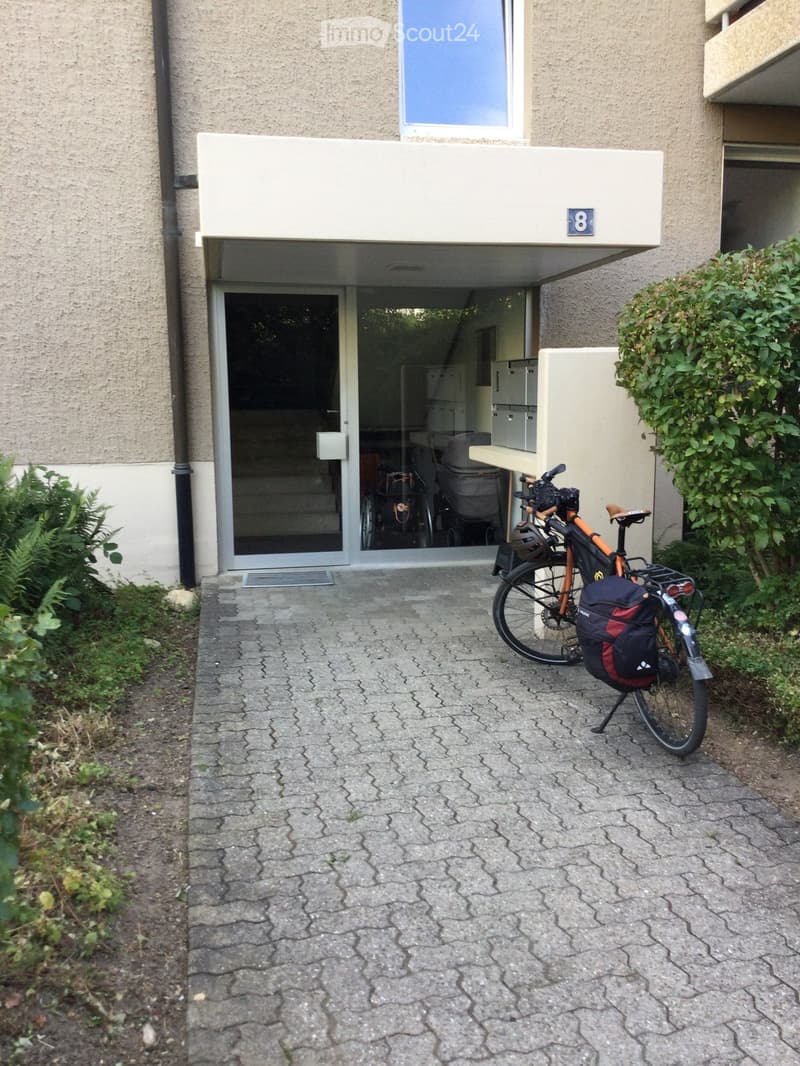 Entrance to a multi-story building with a covered walkway, bicycle parked nearby, and greenery on either side.