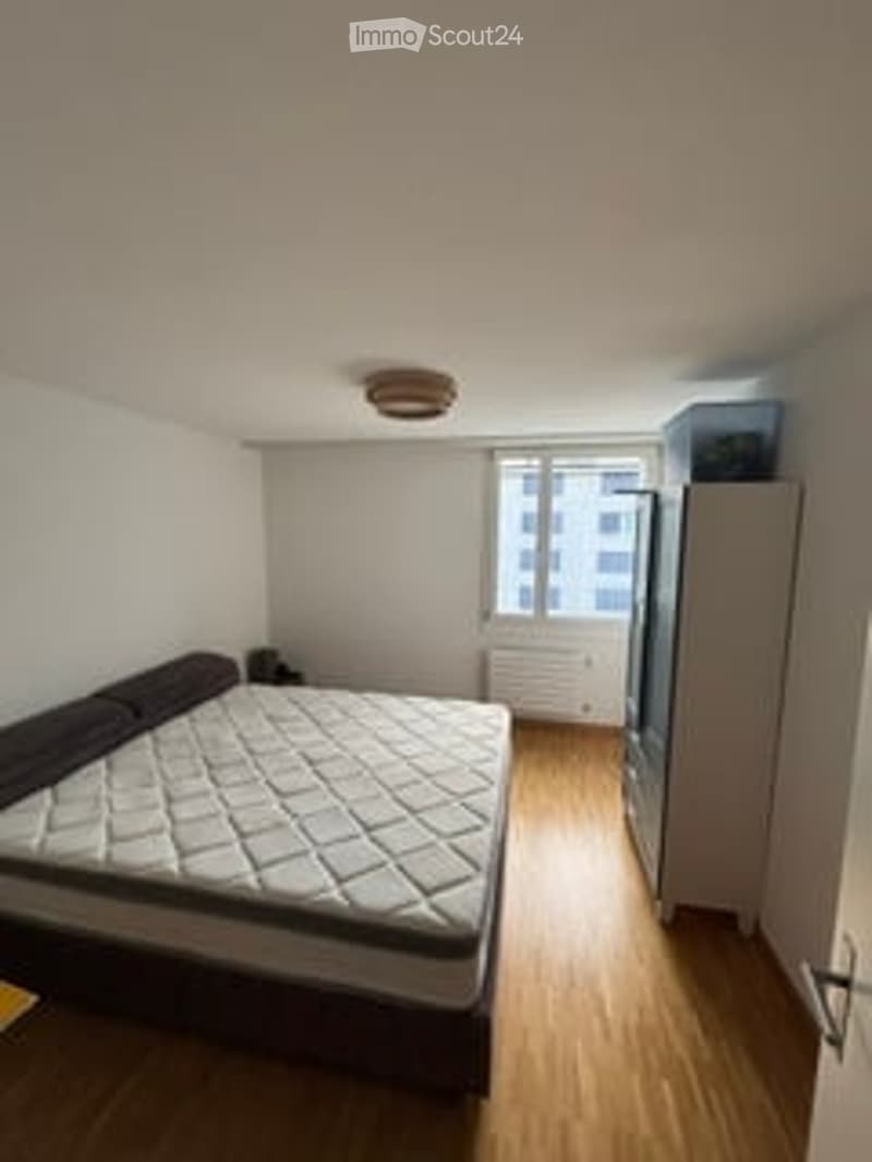 A small, modern bedroom with a large bed, a refrigerator, and a radiator. The room has wooden flooring and large windows with a view of the city.