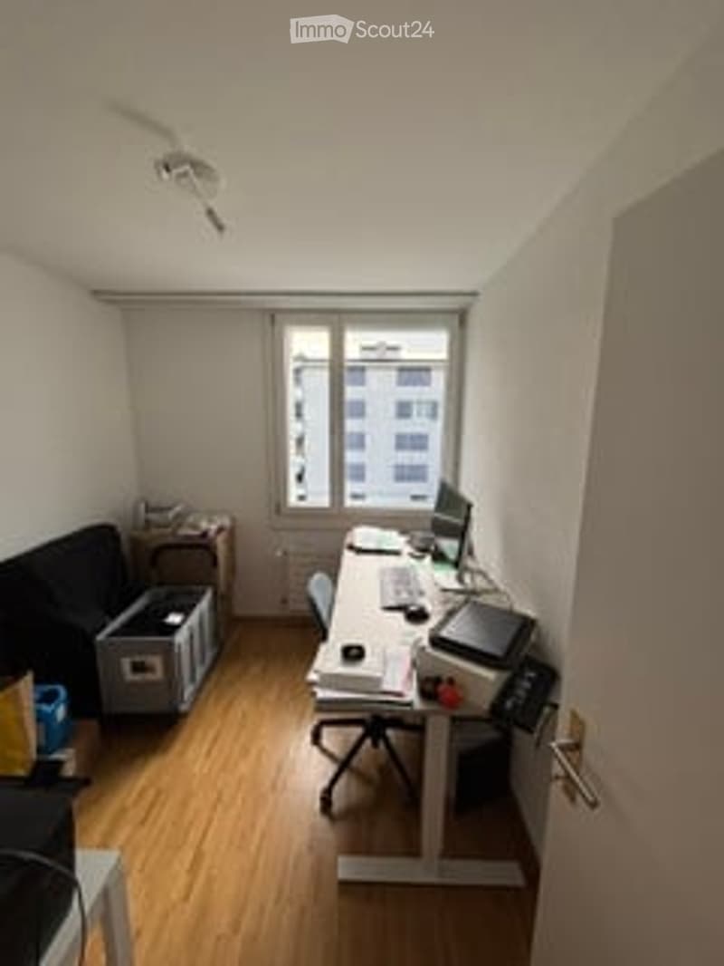 A small, modern office or study room with wooden flooring, white walls, and large windows. The room features a black sofa, a white desk with a laptop, a chair, and a printer. Storage boxes are visible near the sofa.